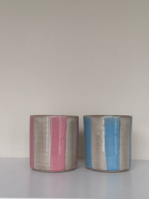 To krus i keramik med lodrette striber, et i rosa og beige og et i blå og beige, placeret på en hvid overflade mod en lys væg.
