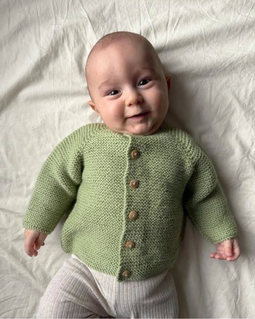 Baby iført lys grøn strikket cardigan og lyse bukser, liggende på hvidt lagen og smilende. (1)