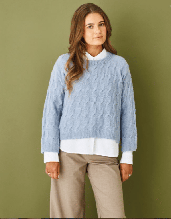 Kvinde iført lyseblå strikket sweater, hvid skjorte og beige bukser, stående foran grøn væg.