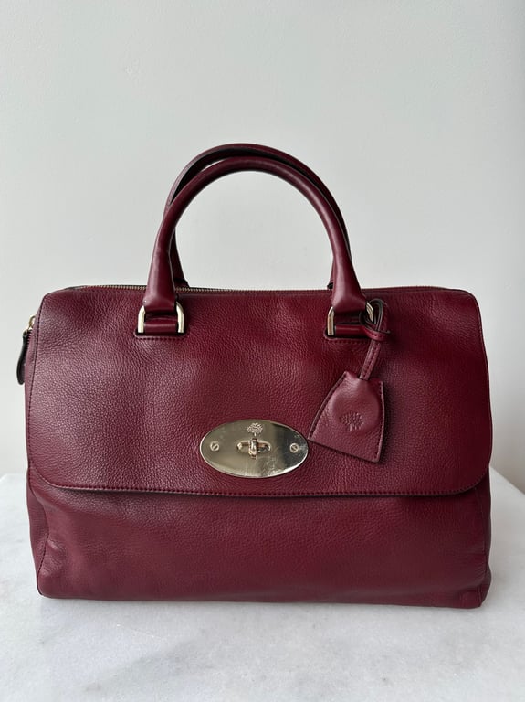 Bordeauxrød lædertaske med håndtag og sølvfarvet lås, placeret på hvid overflade foran en hvid væg.