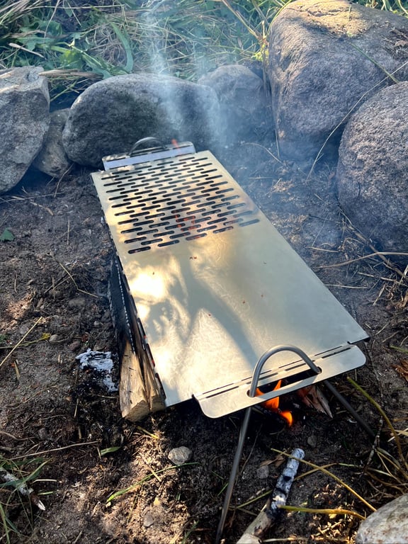 Bærbar metalgrill placeret over et bål, omgivet af sten og natur.