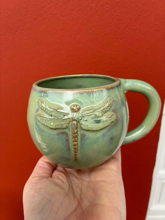 En hånd, der holder en grøn keramikkrus med et indgraveret guldbrynet guldsmedsdesign mod en rød baggrund.