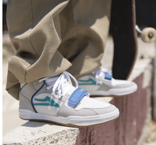 Nærbillede af hvide og blå sneakers på kant, med beige bukser og et skateboard i baggrunden. (1)