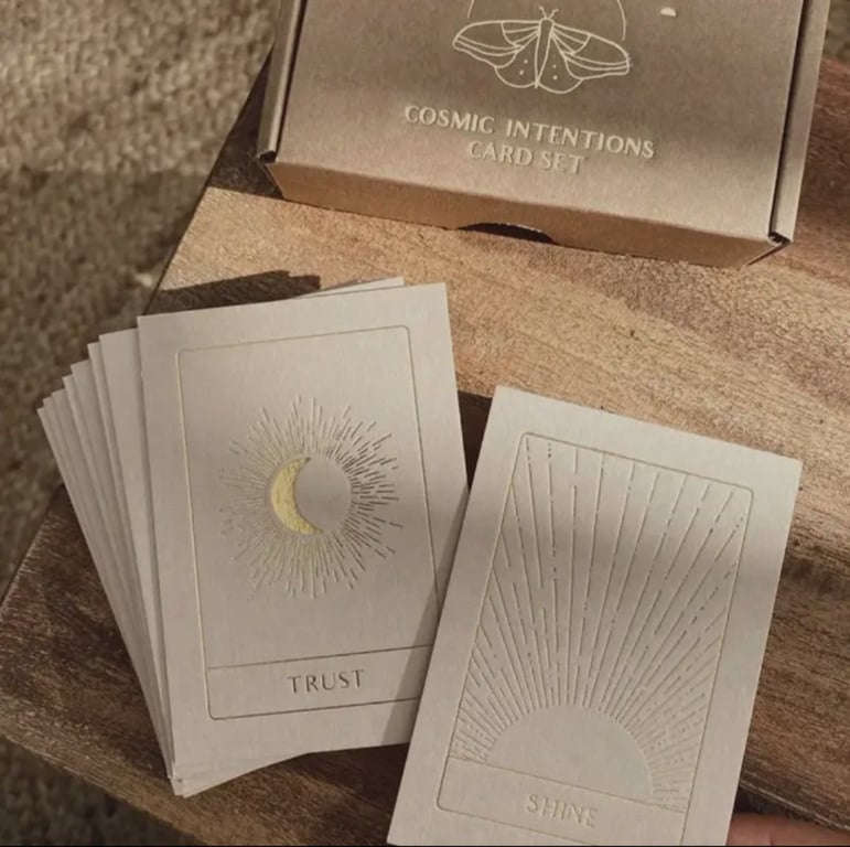 Billedet viser et sæt kort med titlen "Cosmic Intentions Card Set," hvor to kort med motiver af en sol og måne med ordene "TRUST" og "SHINE" ligger på et træbord. (2)