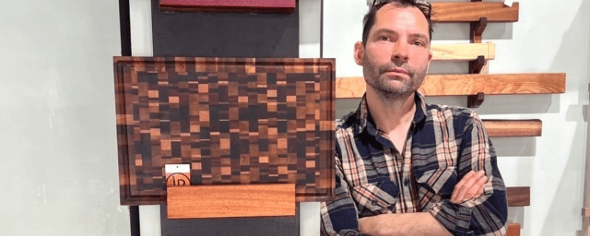 Mand iført ternet skjorte med armene over kors står ved siden af et mønster af forskellige træplader monteret på væggen.