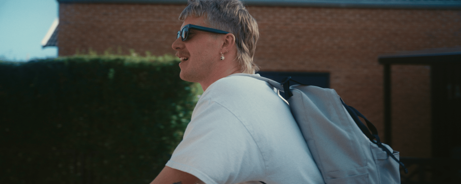 Mand med solbriller, hvid t-shirt og rygsæk cykler forbi et hus med murstensfacade på en solrig dag.