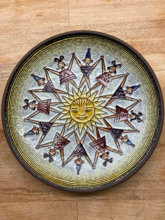 Rund keramikplade med et solmotiv i midten omgivet af figurer, der holder hinandens hænder i en cirkel, på en træflade. (2)
