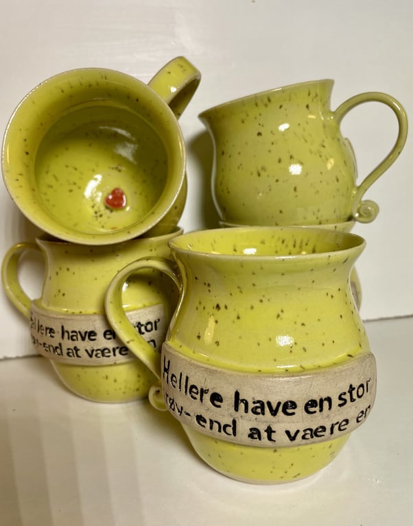 Gulbrune keramikkrus med hank og sort tekst på beige bånd, en af kopperne har en lille rød hjertefigur indeni.