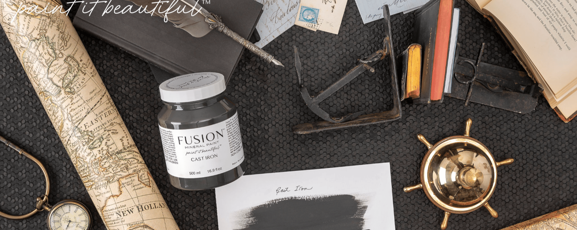 En flaske Fusion mineralmaling i farven Cast Iron omgivet af en antik globus, åbne bøger, håndskrevne breve, en lommeur, en lille guld styrerem og en sort notesbog med en fjerpen på en sort baggrund.