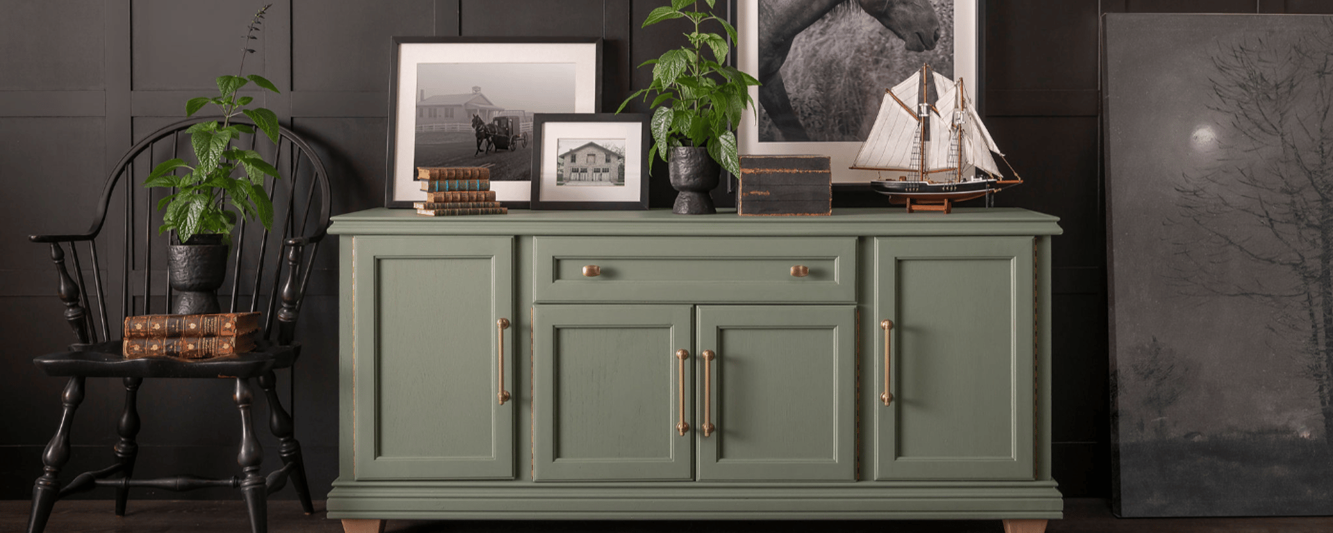 Grønt skænkbord med messinghåndtag, dekoreret med planter, gamle bøger, et modelskib og indrammede sort-hvide fotografier, placeret mod en mørk væg ved siden af en sort træstol med planter og bøger.