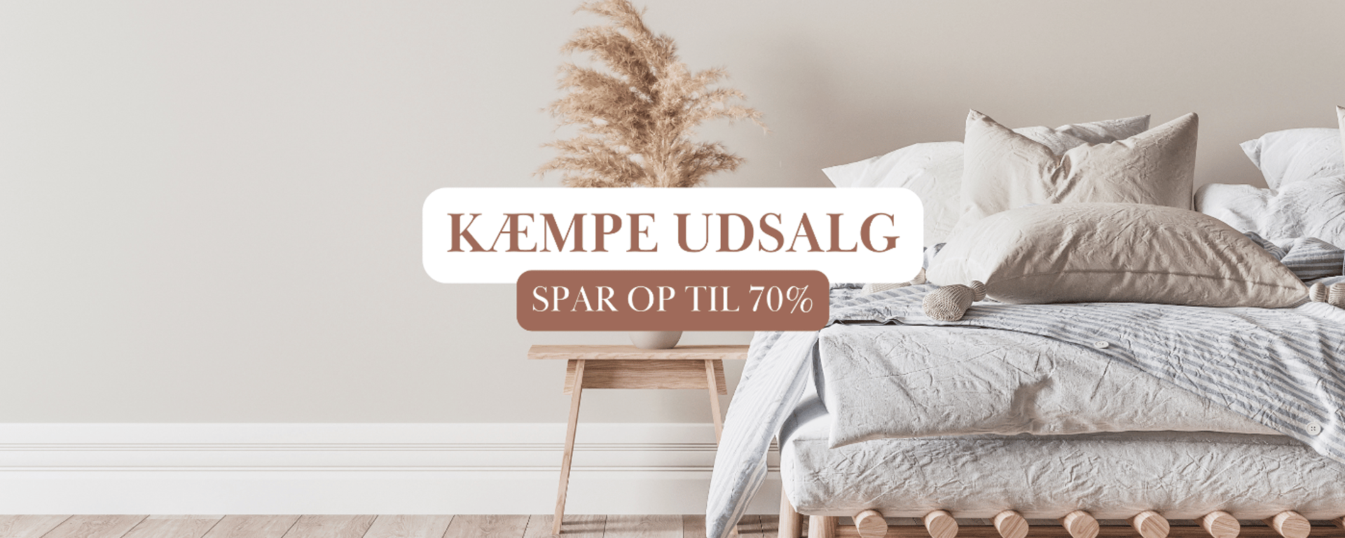 Soveværelse med beige sengetøj og puder, søjlebord med tørrede blomster og tekst om kæmpe udsalg med op til 70% rabat.