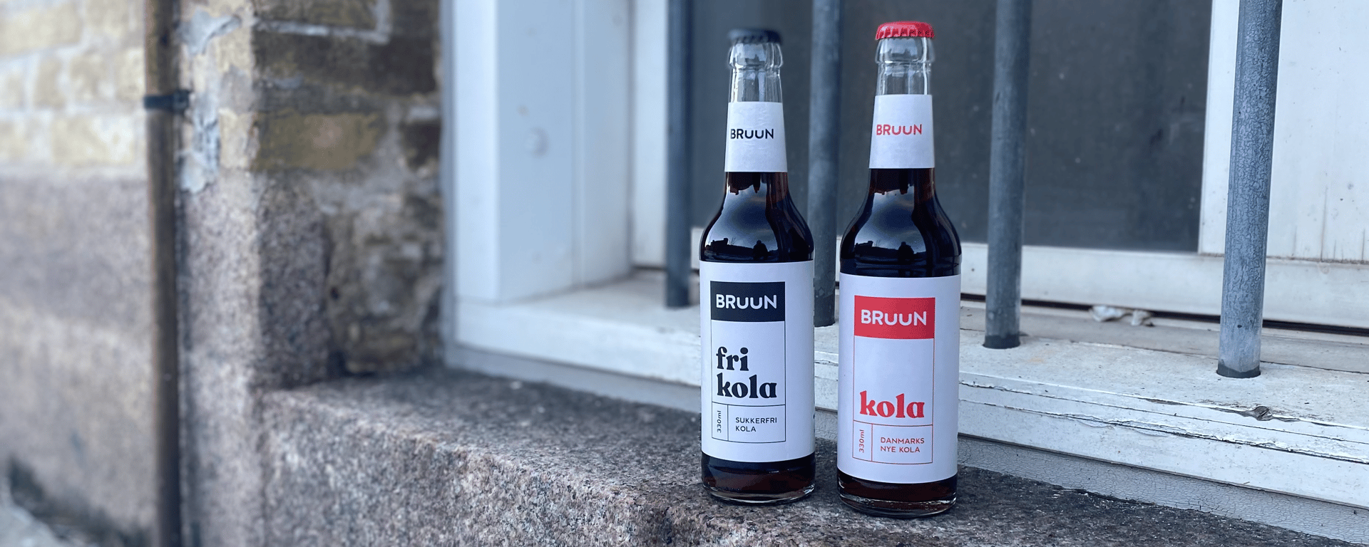 To flasker med cola, en med mærket "fri kola" og en med mærket "kola", står på en sten ved en bygning med en dør i baggrunden.