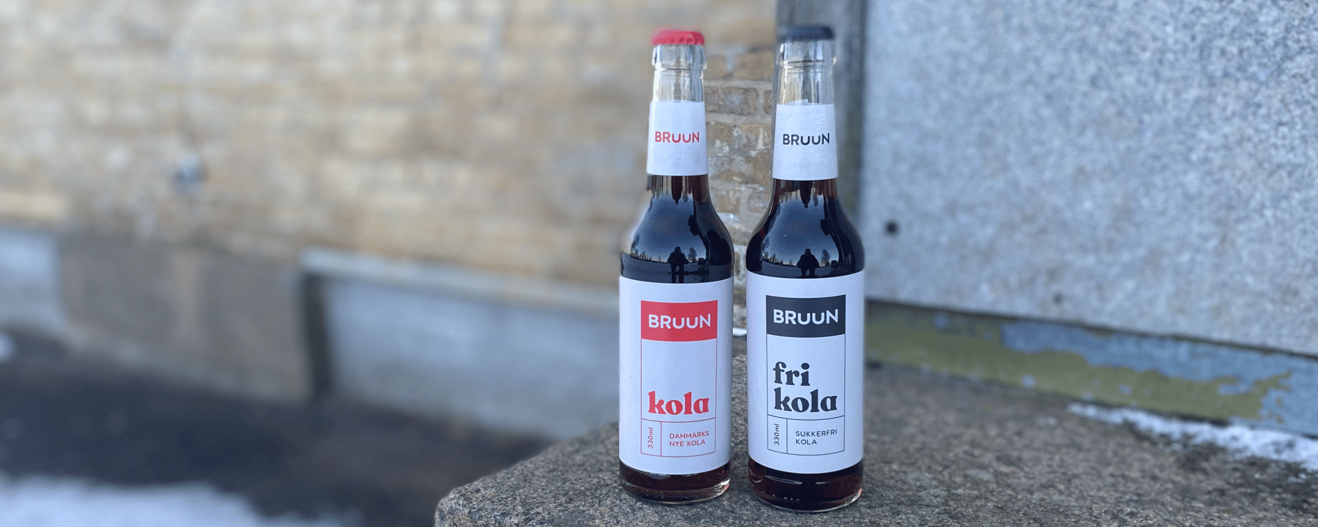 To flasker bruun kola, én med sukkerfri variant, placeret på en stenet overflade udendørs foran en væg.