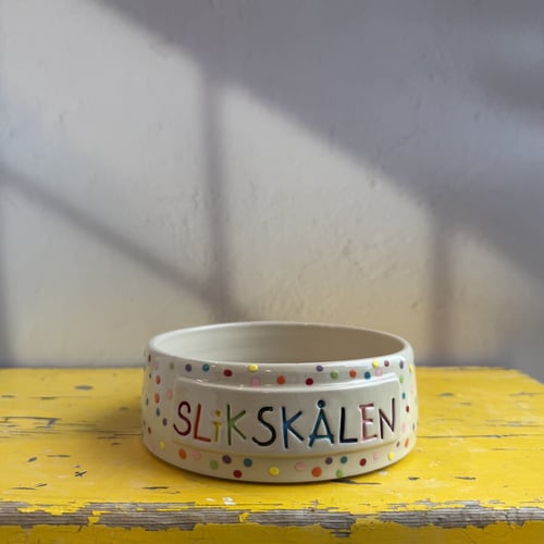 En hvid keramikskål med farverige prikker og teksten "SLIKSKÅLEN" i forskellige farver, placeret på et slidt gult bord med skygger på væggen bagved. (1)