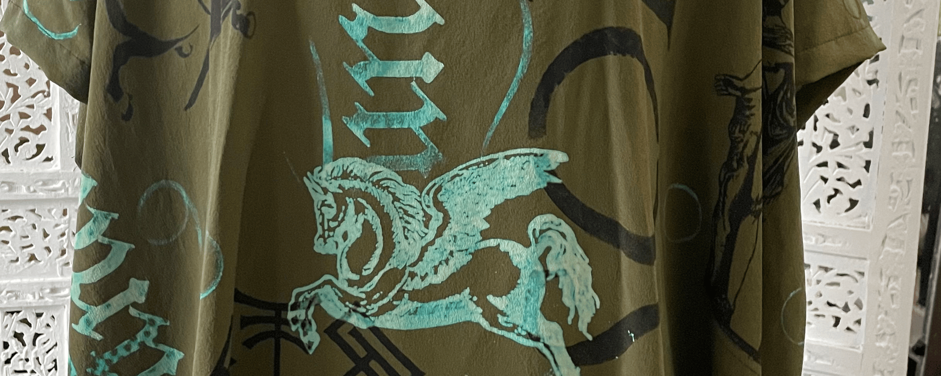 T-shirt med grøn base og lys turkis tryk af en bevinget hest samt gotisk tekst og andre dekorative mønstre.