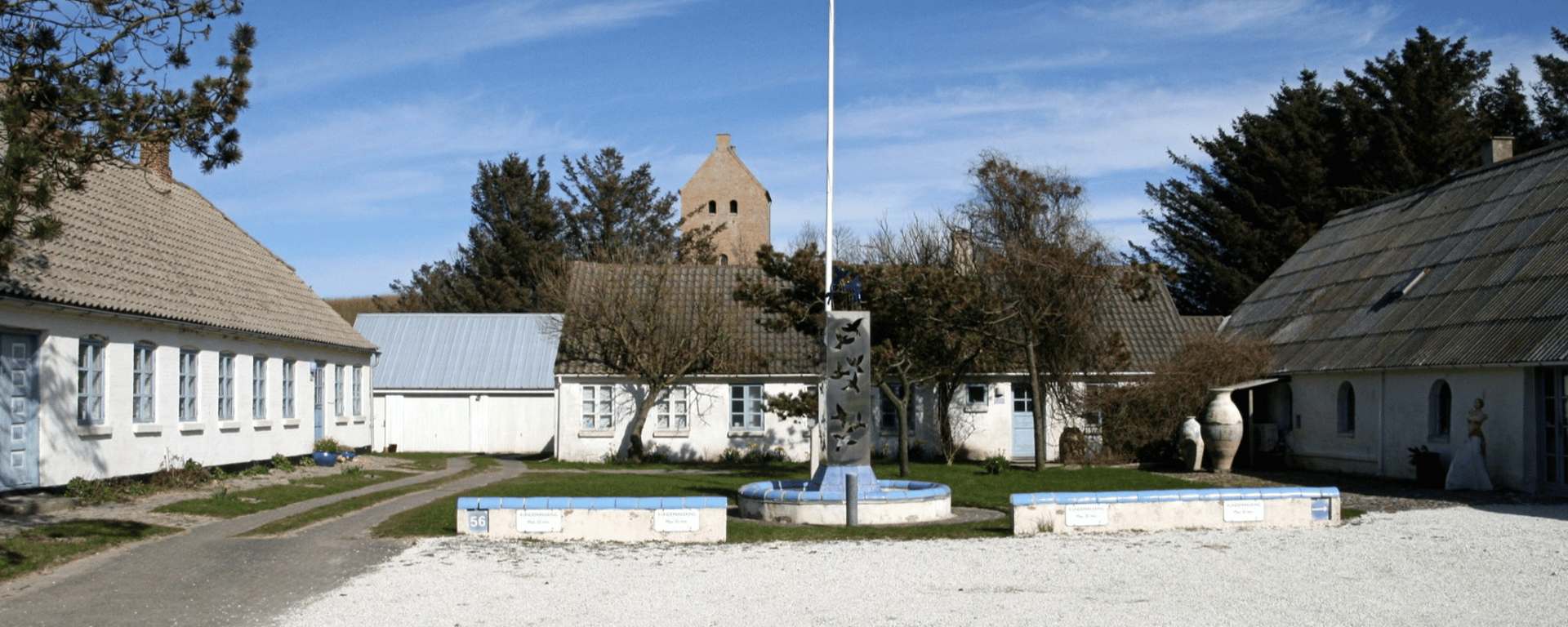 Gårdmiljø med hvide bygninger og grå tage omkring en cirkulær bænk og en flagstang, med træer og et kirketårn i baggrunden under en klar blå himmel.