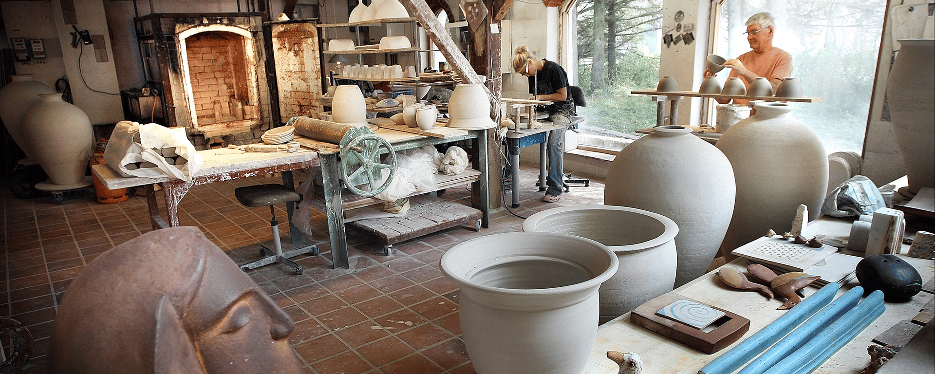 Keramikerværksted med store lerkrukker, skulpturer og to personer, der arbejder med keramiske emner ved store vinduer med udsigt til træer.