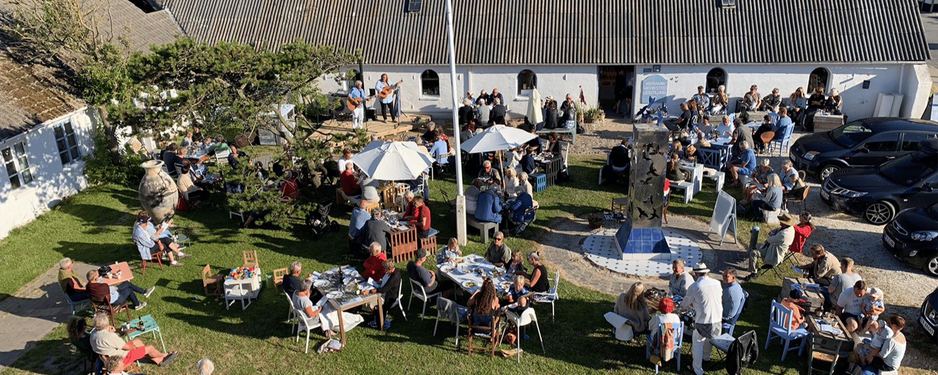 Udendørs arrangement i en gårdhave med mange mennesker, der sidder ved borde og nyder musik fra to guitarister ved en hvid bygning.