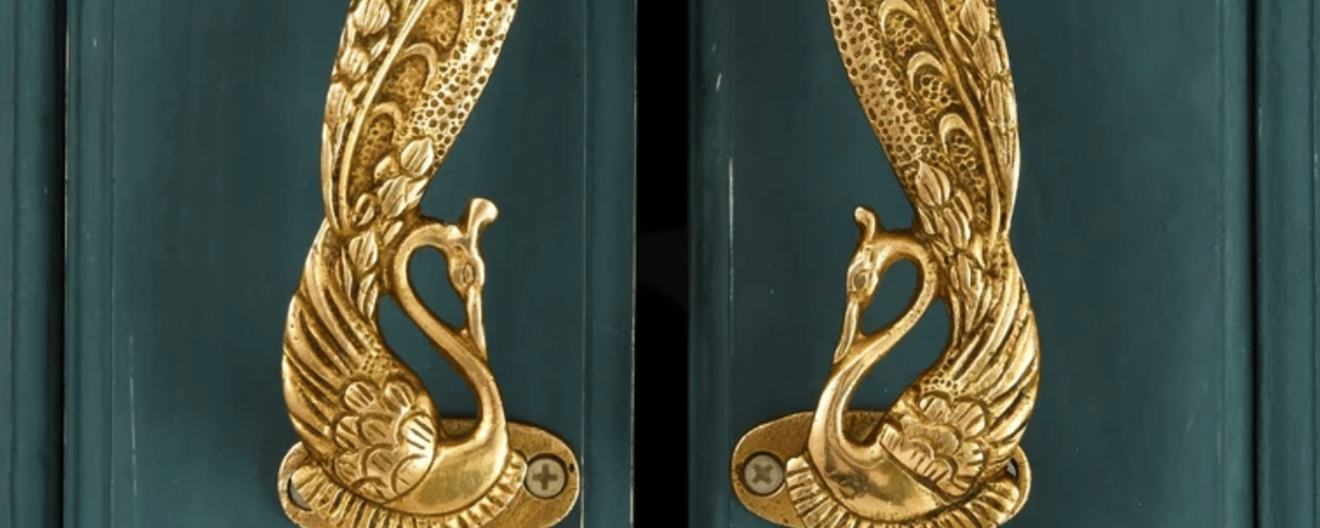 To dørhåndtag i messing formet som detaljerede svaner med oprejste vinger på en mørk grøn dør.
