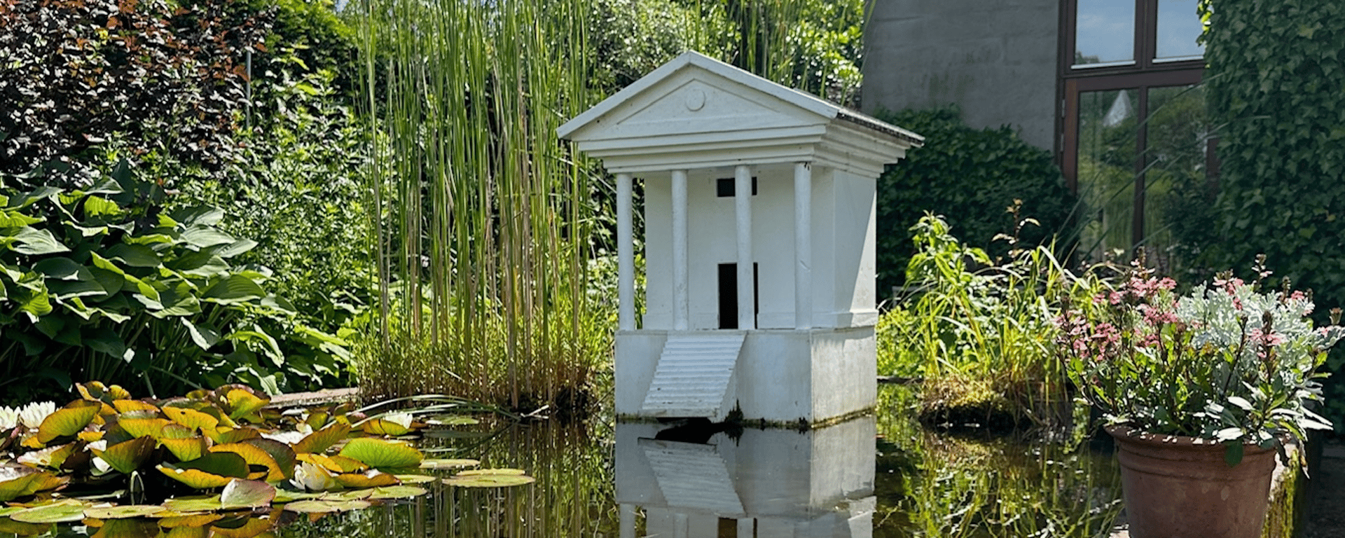 Model af et hvidt tempel med søjler, der står på en lille dam omgivet af planter og blomster i en have.