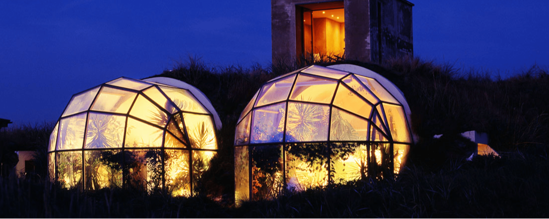 To gennemsigtige geodætiske kupler oplyst indefra om aftenen, omgivet af vegetation og en bygning i baggrunden.