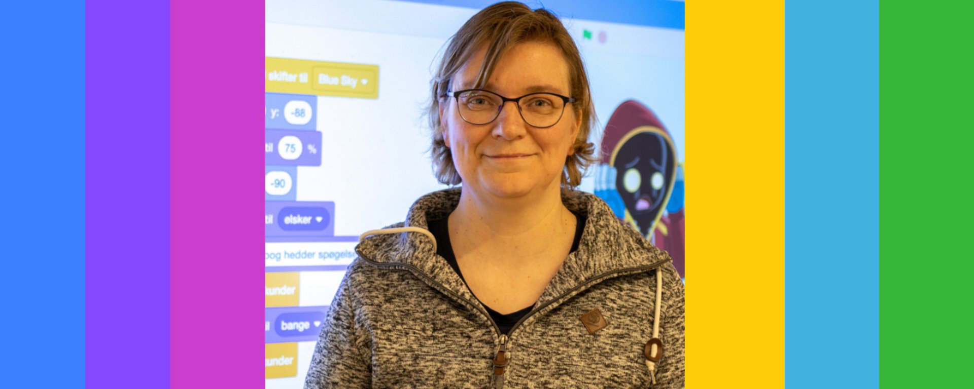 Kvinde med briller iført grå hættetrøje står foran en skærm med programmeringsblokke og animeret figur.