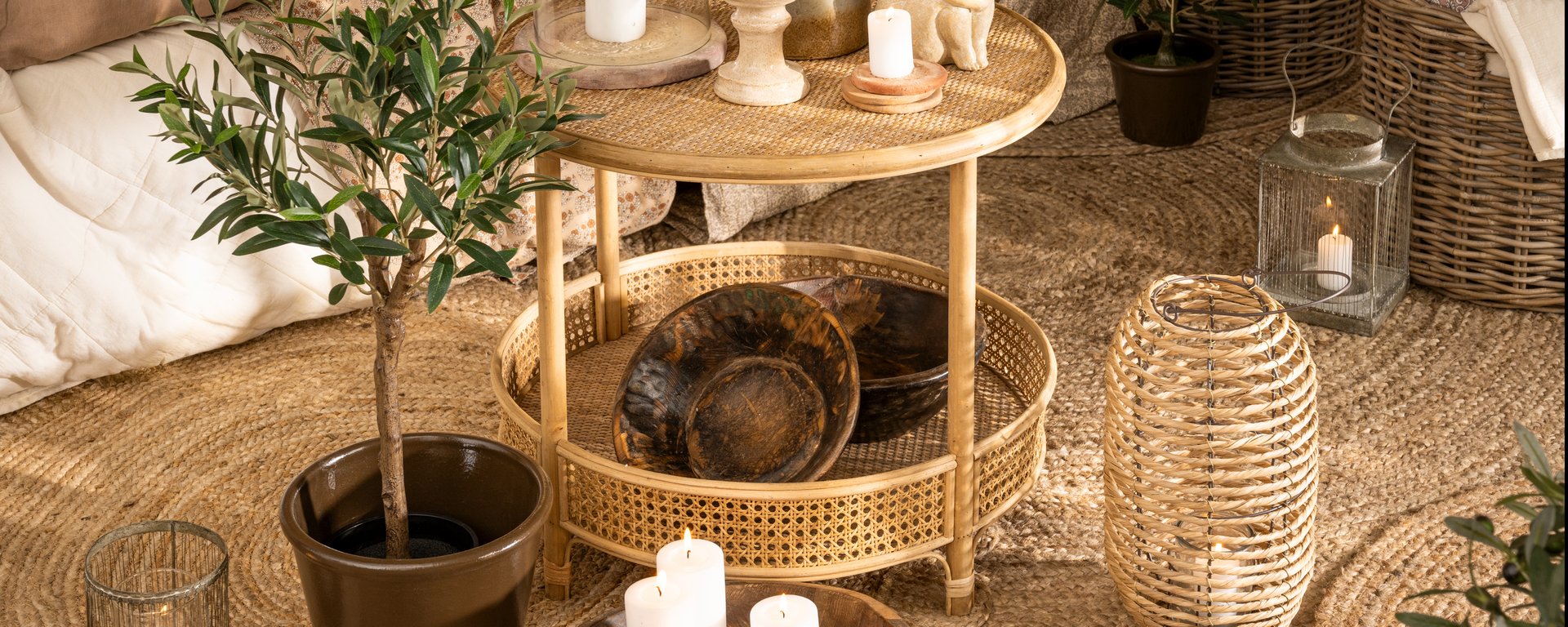 Lille rundt fletbord med to mørke skåle, levende stearinlys og potteplante på en rund tæppebund i naturmaterialer, omgivet af lanterner og kurve.