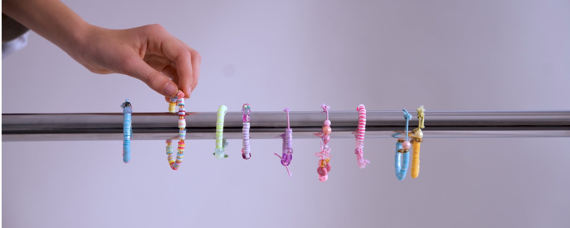 En person placerer eller tager et lyst, farverigt armbånd med perler fra en række med otte forskellige farverige armbånd, der hænger på en metalstang mod en lysegrå baggrund.