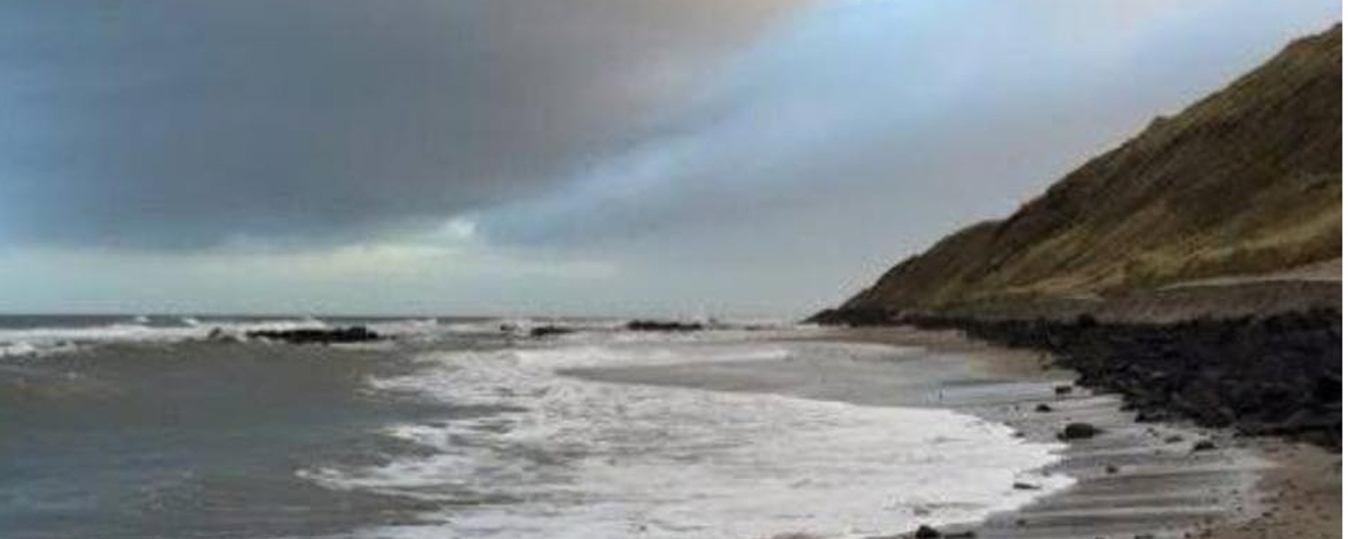 Strand ved kysten med skum på vandet, skrånende græsklædt bakke og overskyet himmel.