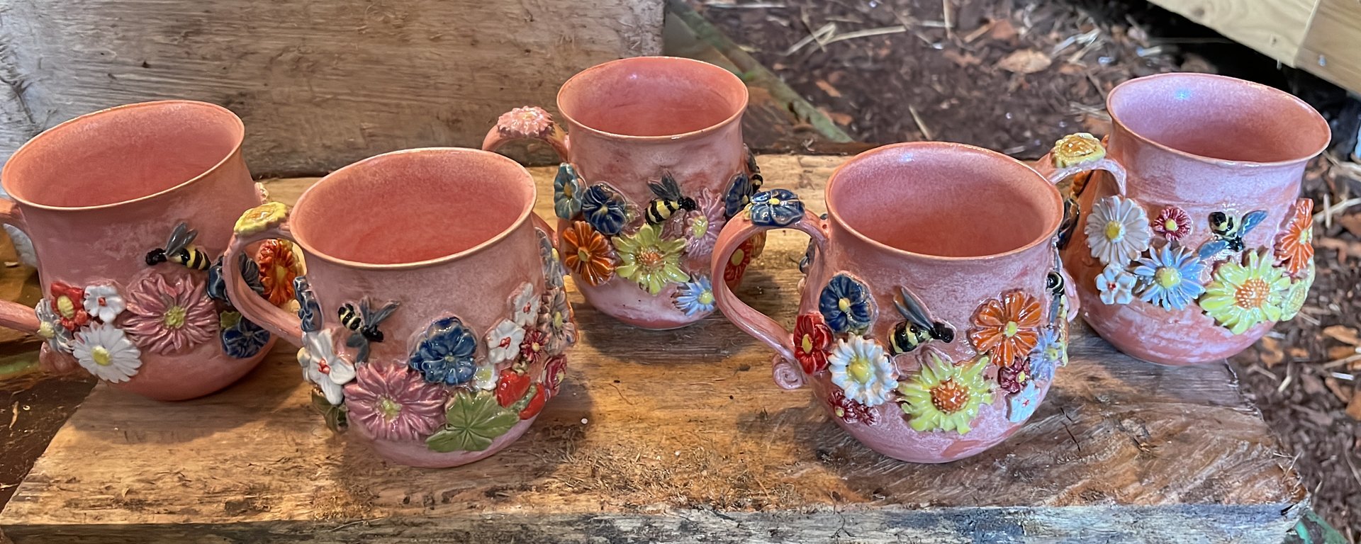 Fem lyserøde keramikmugger med farverige blomster og bier som dekoration, placeret på en rustik træflade.