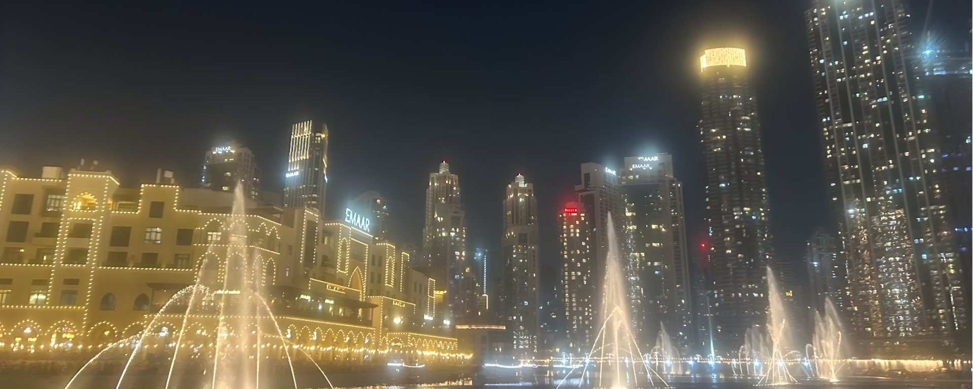 Natteligt bybillede med oplyste højhuse, fontæner med vandstråler og bygninger dekoreret med lys.