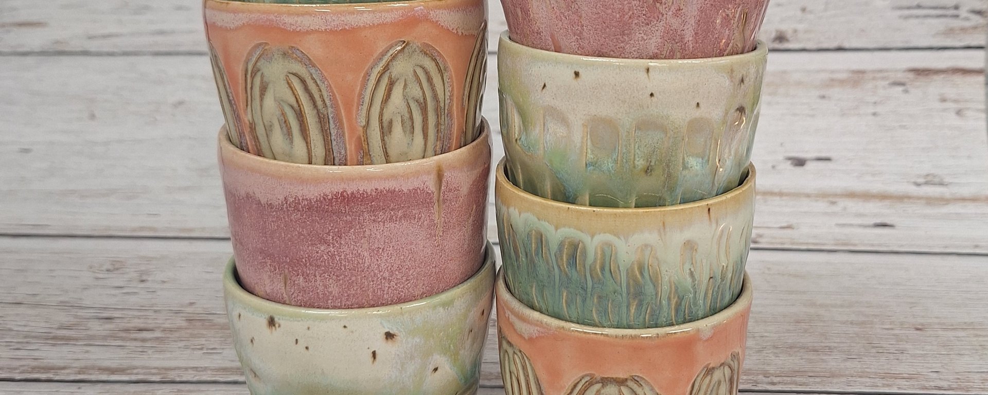 To stakke keramik kopper i forskellige pastelfarver med abstrakte mønstre, placeret på et lyst træbord og baggrund.