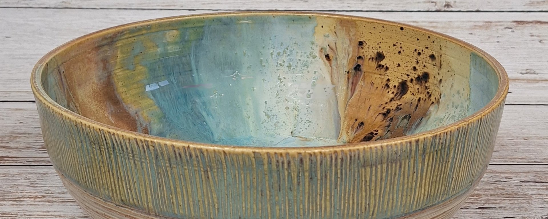 Skål i keramik med blå, grønne og brune farver og tekstureret yderside, placeret på et lyst træbord.