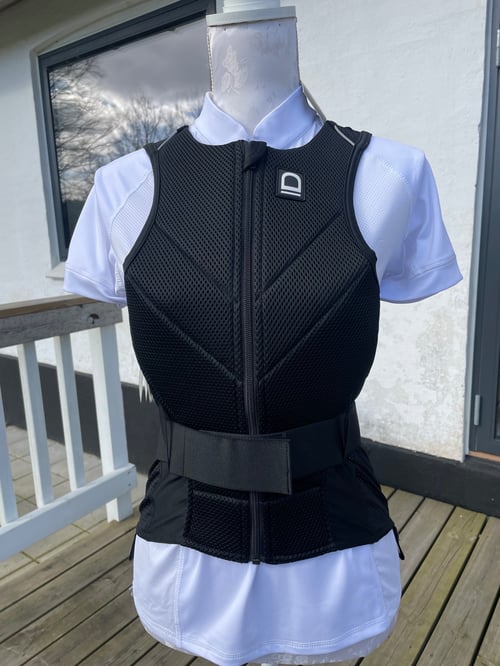 Sort beskyttelsesvest med lynlås og bred velcrobælte, vist på en mannequin iført en hvid skjorte, stående på en træterrasse foran et hvidt hus. (1)