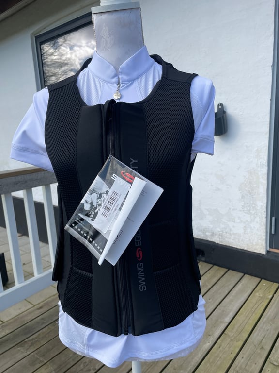 Mannequin iført hvid ridebluse og sort sikkerhedsvest med tags, placeret på en træterrasse udenfor.