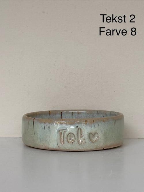 Keramisk skål med teksten "Tak" og et hjerte præget på siden, mod en beige væg med ordene "Tekst 2 Farve 8" øverst til højre. (1)