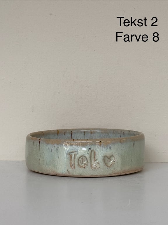 Keramisk skål med teksten "Tak" og et hjerte præget på siden, mod en beige væg med ordene "Tekst 2 Farve 8" øverst til højre.