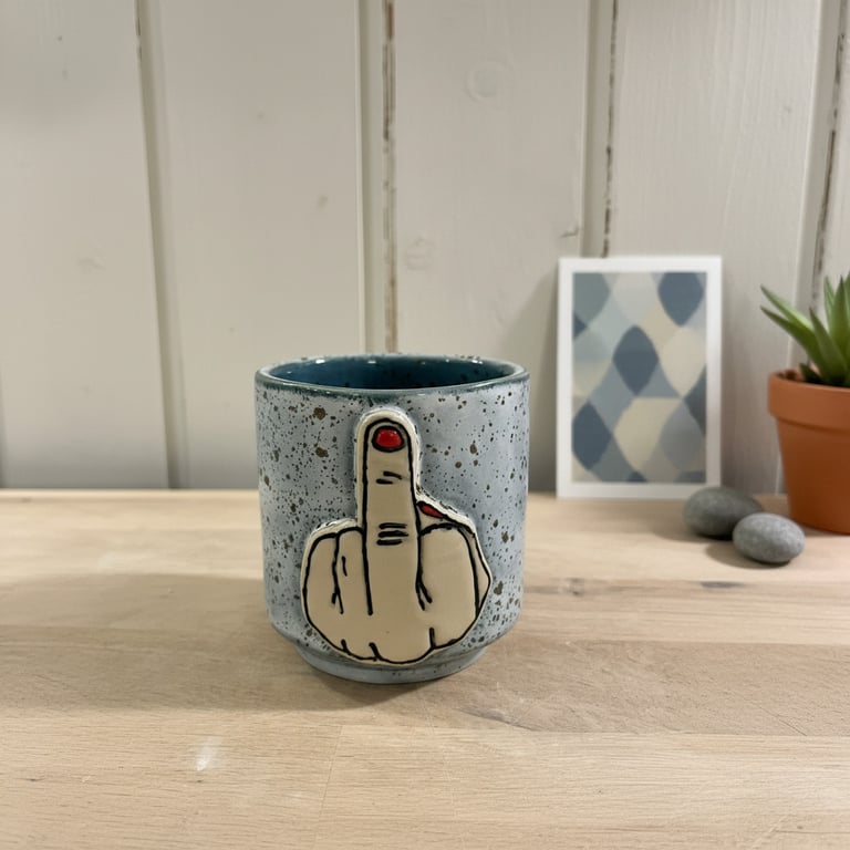 Grå keramik kop med tegning af en hånd, der viser langemand med rød neglelak, placeret på et lyst træbord med plante og billedramme i baggrunden.