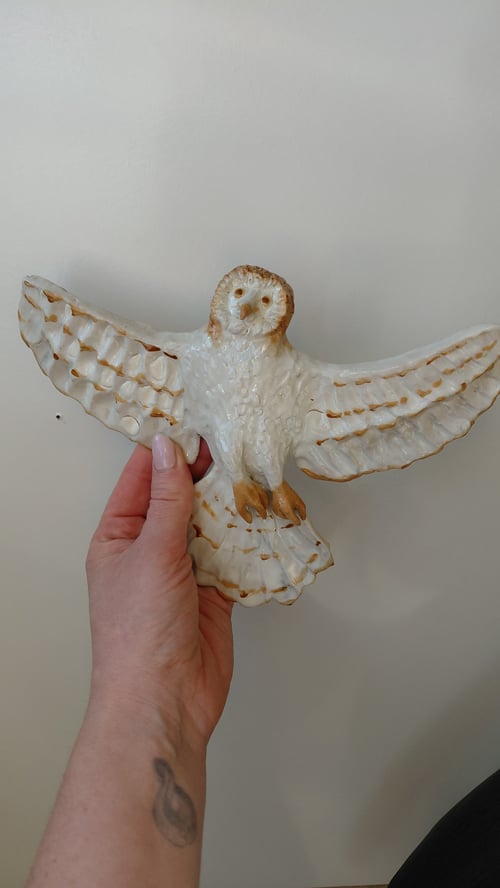 Hånd, der holder en hvid keramisk uglefigur med udstrakte vinger mod en lys baggrund. (1)