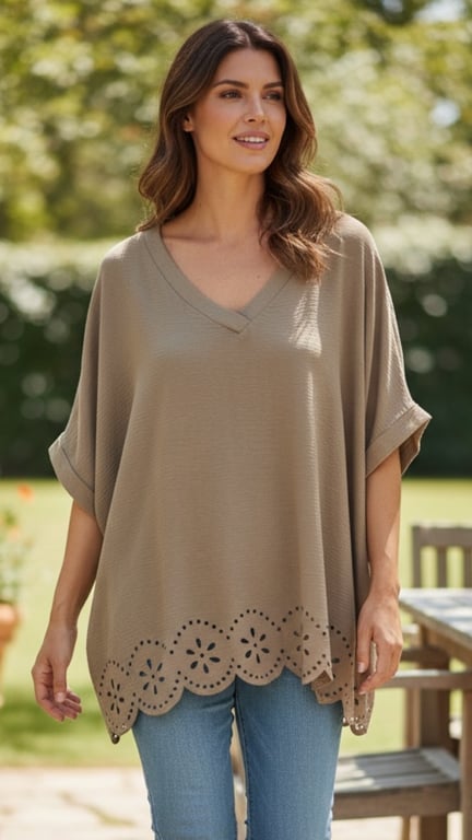Kvinde iført beige oversized bluse med V-hals og blomsterudskæringer i kanten, stående i en solrig have.