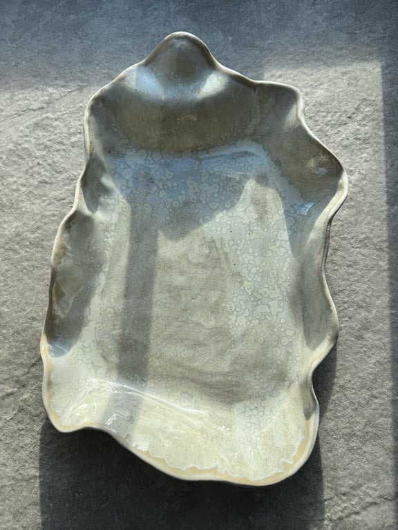 Uregelmæssigt formet, håndlavet keramisk fad med glaseret, lys overflade og bølgede kanter, placeret på en grå tekstilbaggrund i naturligt lys.