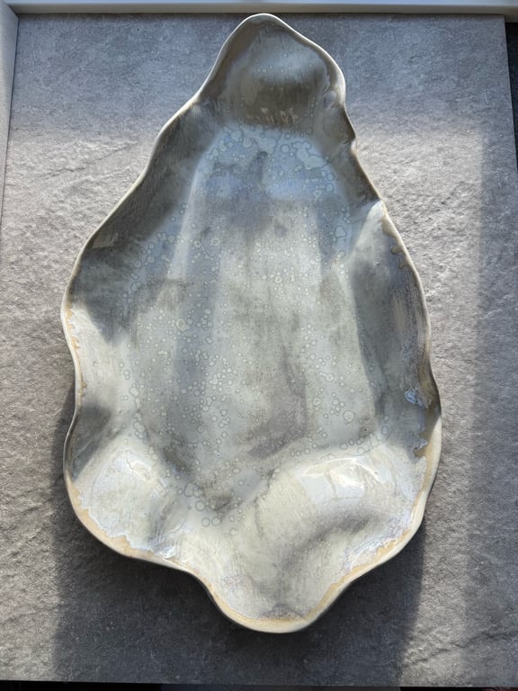 Unik, håndlavet keramikskål med uregelmæssig form og grålig glasur med små prikker, placeret på en grå overflade.