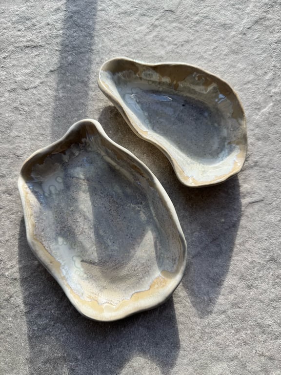 To keramiske fade med uregelmæssige former i grå og beige nuancer, placeret på en lys overflade med sollys og skygger.