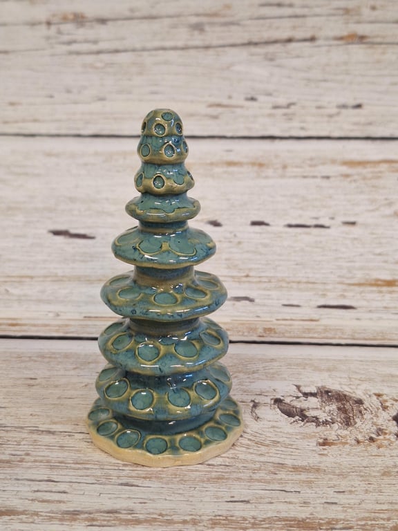 Keramisk, blågrøn skulptur med cirkelformede mønstre, formet som et tårn, placeret på et træbord.