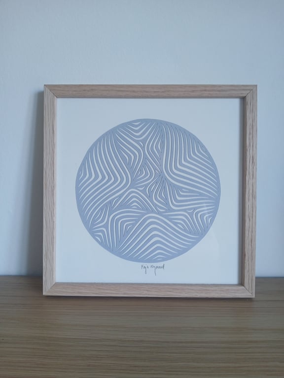 Indrammet abstrakt kunstværk med bølgende, hvide linjer i en cirkel på grå baggrund, placeret på et træbord mod en lyseblå væg.