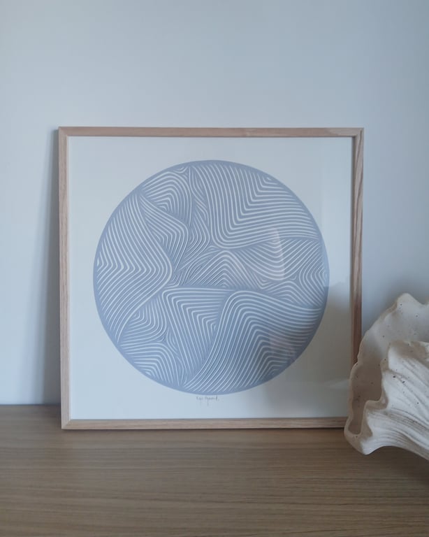 Indrammet kunstprint med abstrakt cirkelformet mønster i blå og hvide linjer, placeret på et træbord ved siden af en stor hvid muslingeskal.