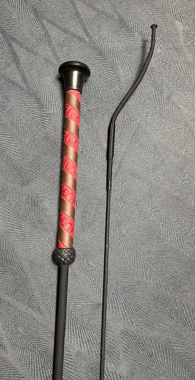 To sort riding crop med brun og rød håndtag, lagt på gråt mønstret stof.