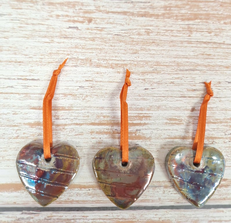 Tre keramiske hjerteformede pynt med orange bånd, arrangeret på en lys trægulvbaggrund.
