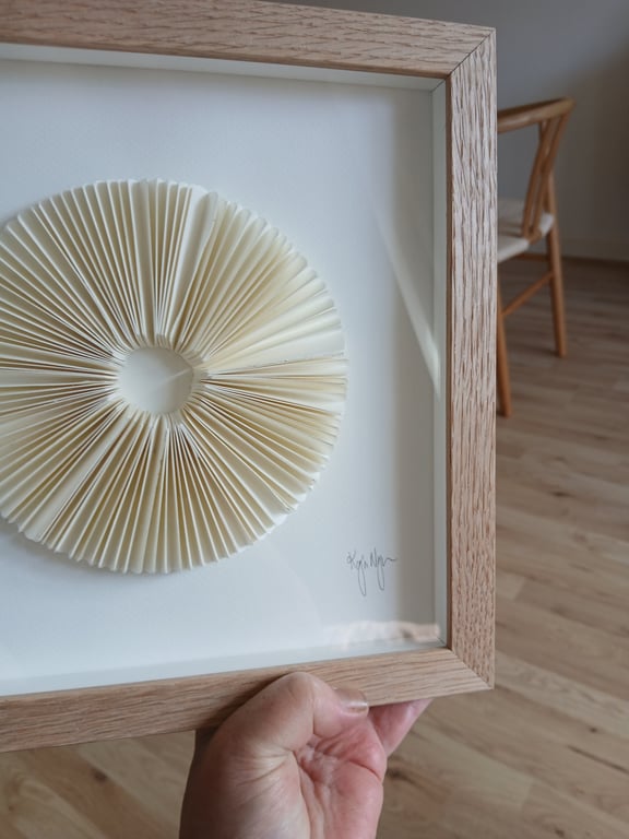 Hånd, der holder en indrammet kunstværk med et foldet, cirkulært papirdesign, i et rum med træstol og trægulv.