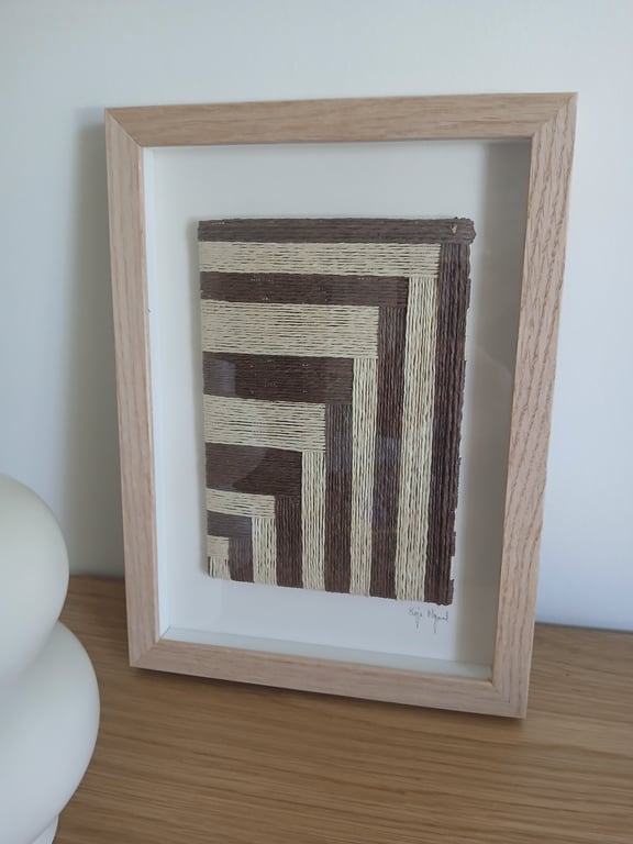 Indrammet kunstværk med geometrisk mønster i brune og beige nuancer på en hvid baggrund, placeret på et træbord ved siden af en hvid skulptur.